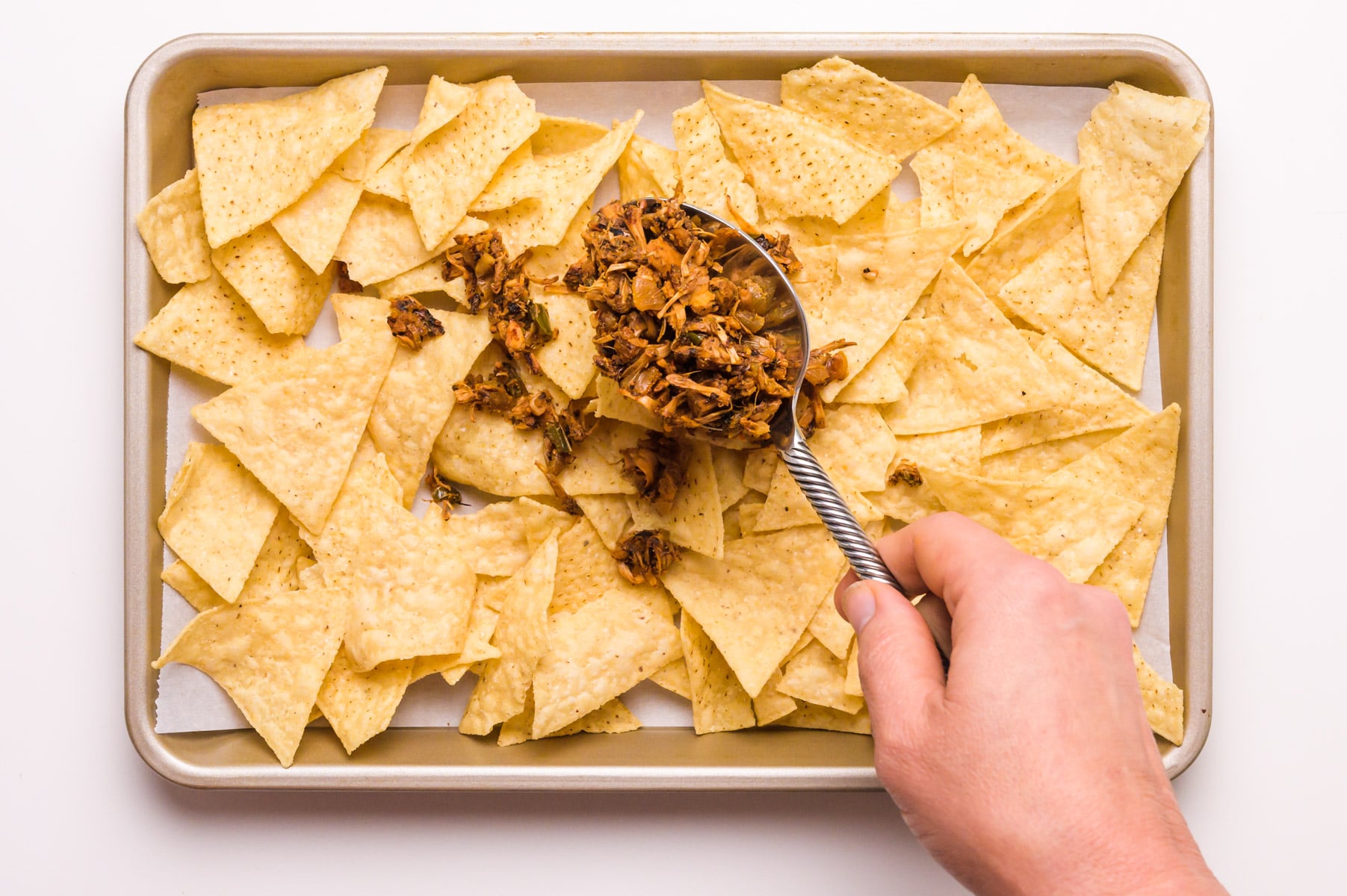 A hand holds a spoon dispensing jackfruit carnitas onto a pan filled with tortilla chips.