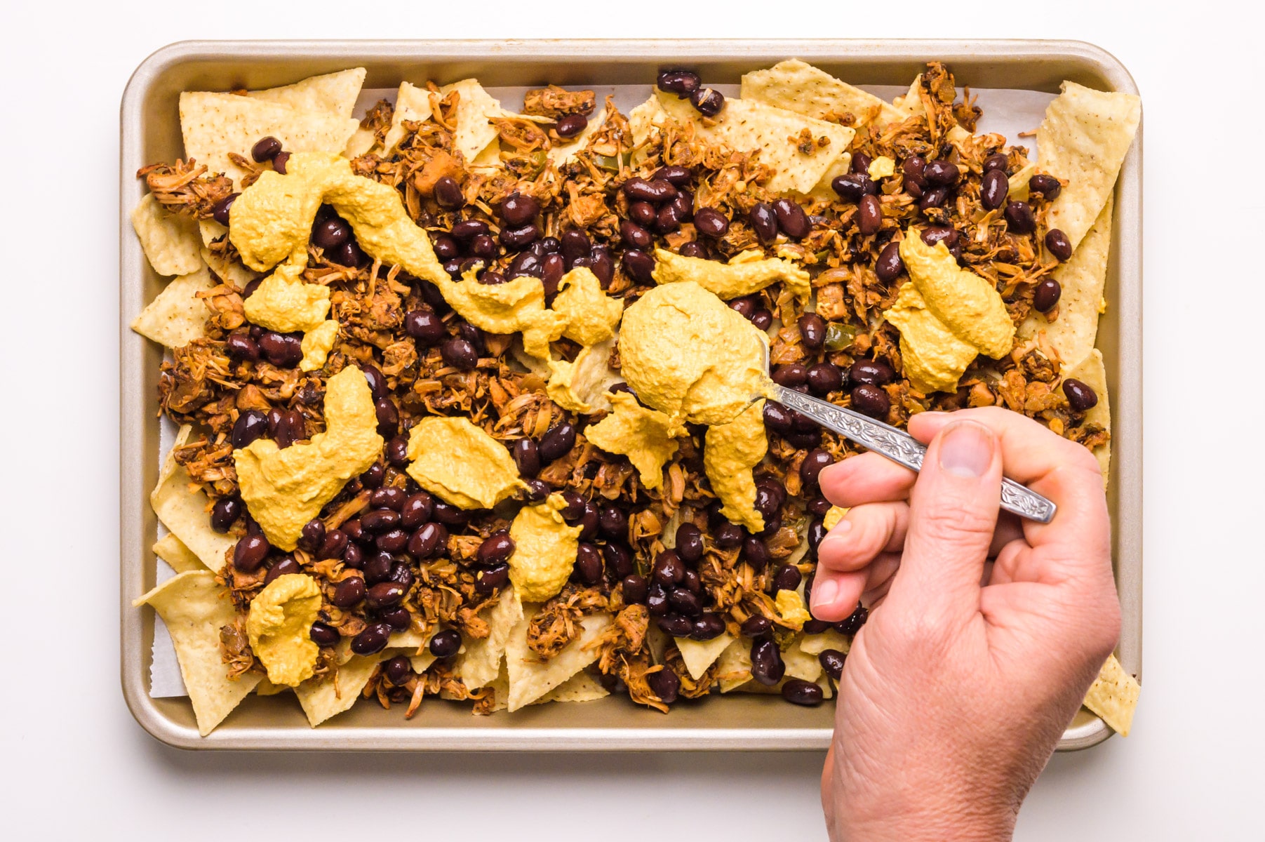 A hand holds a spoon distributing vegan nacho cheese on top of a pan of nachos.