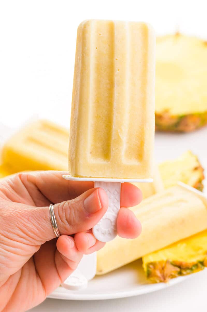 One hand holds a pineapple popsicle in front of more popsicles and fresh pineapple in the background.