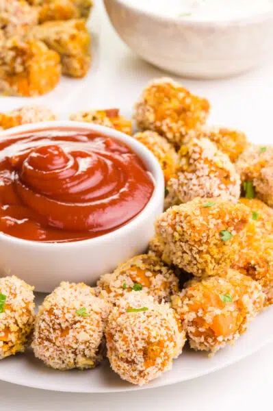 A plate of tater tots sits around a bowl of ketchup. There is another bowl of dip and tater tots in the background.