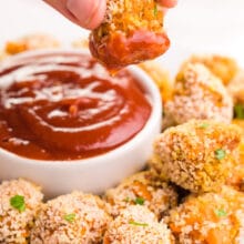 A hand dangles a sweet potato tater tot dipped in ketchup, more tater tots and a bowl of ketchup on a plate.