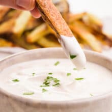 A hand holds french fries in a bowl of vegan aioli.