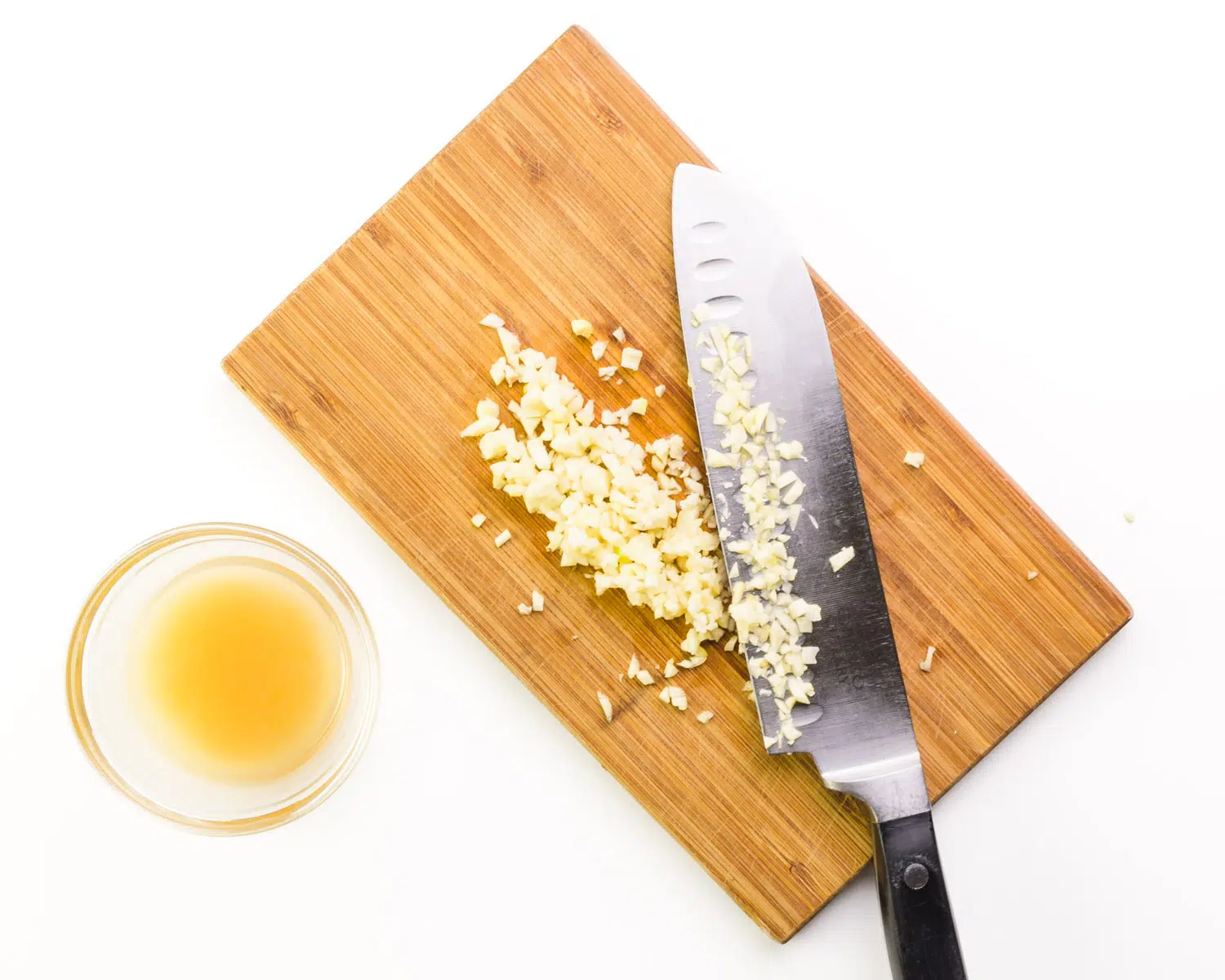 A chef's knife is on a cutting board with minced garlic. There is a bowl of lemon juice next to it.