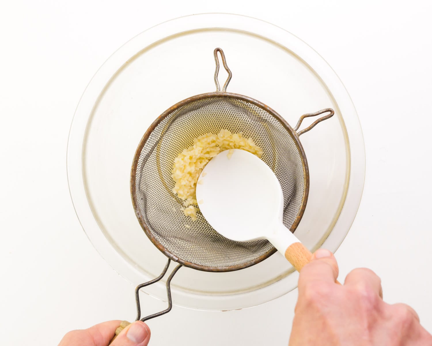 Garlic is being pressed with a spatula into a fine mesh strainer positioned over a glass bowl.