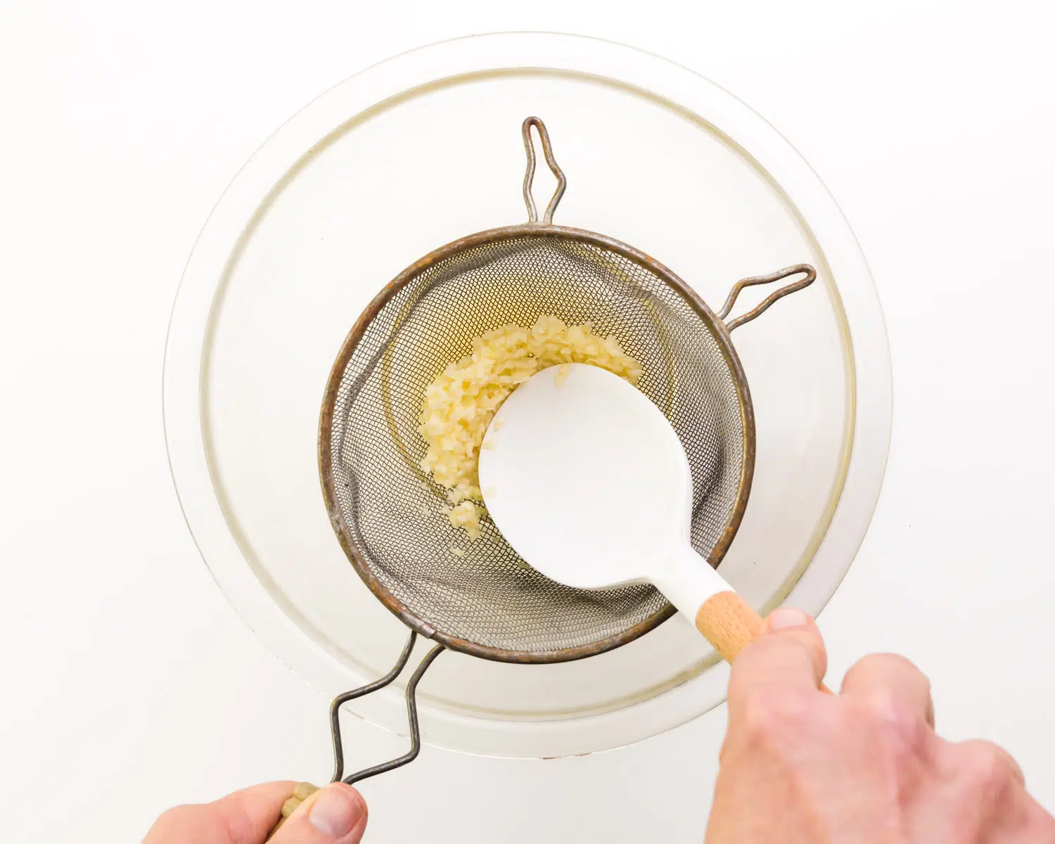 Garlic is being pressed with a spatula into a fine mesh strainer positioned over a glass bowl.