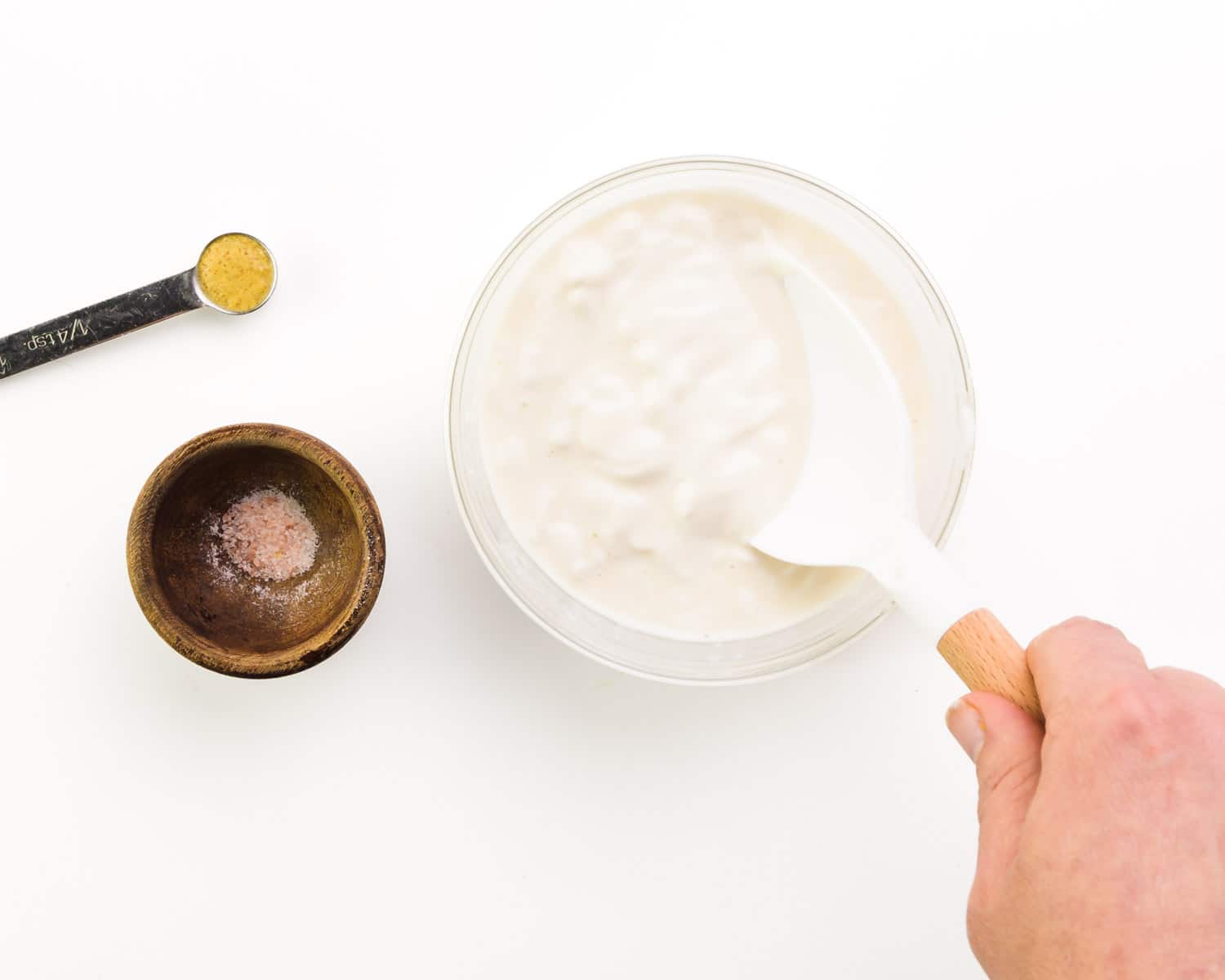 A hand holds a spatula, stirring vegan aioli in a bowl.  Have a measuring spoon with brown mustard and a bowl of sea salt next to it.