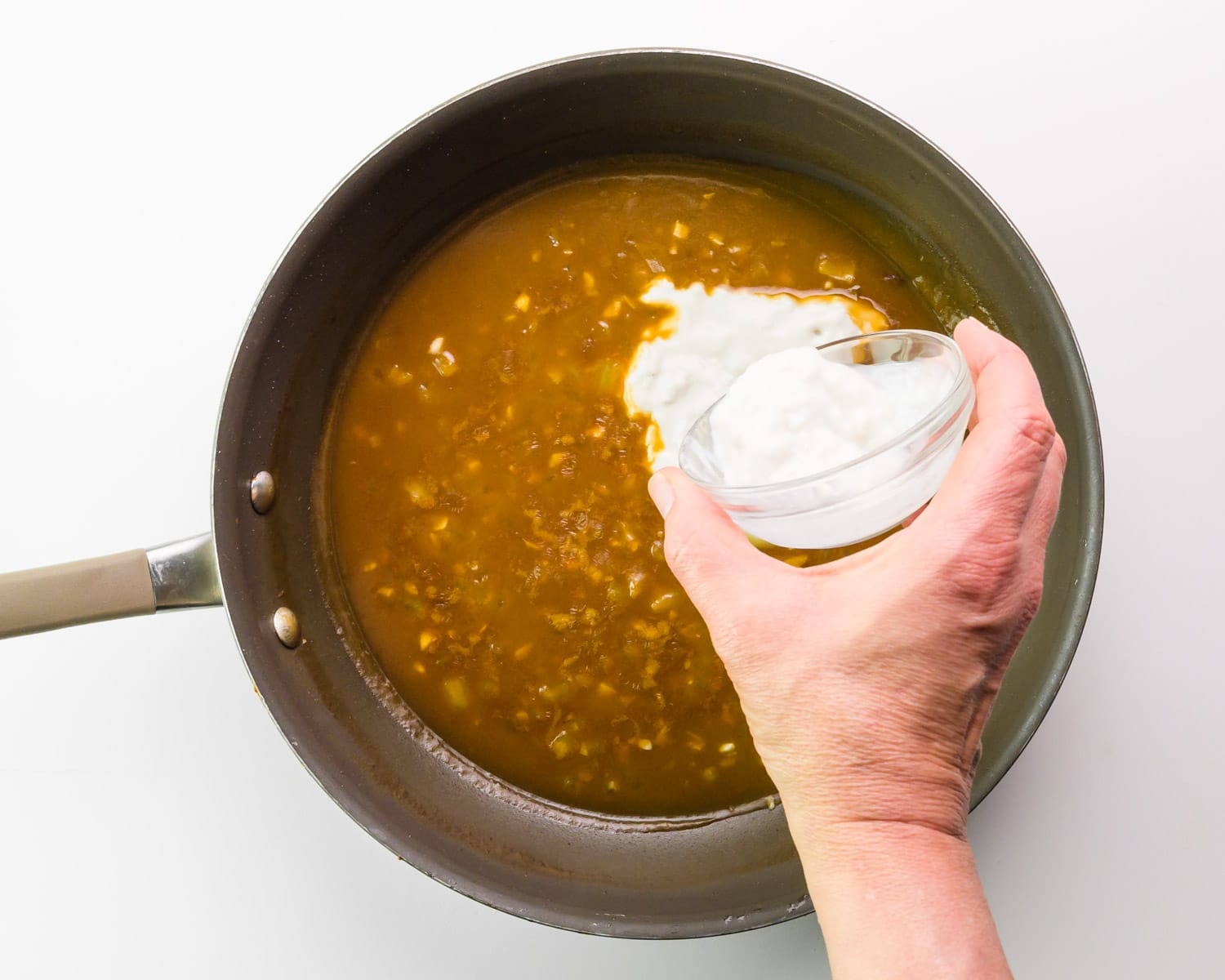 A hand holds a bowl of vegan sour cream, pouring it into a skillet with a brown sauce.
