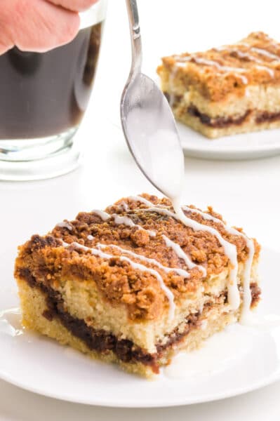 A spoon drizzles icing over a slice of vegan coffee cake. Another slice is in the back beside a cup of coffee.