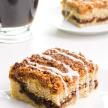 A slice of coffee cake on a plate sits in front of another slice and a cup of coffee.