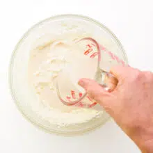 A hand holds a measuring glass full of plant-based milk pouring it into a bowl with whipped vegan butter.