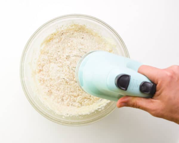 A hand holds a blue mixer, beating batter in a glass bowl.