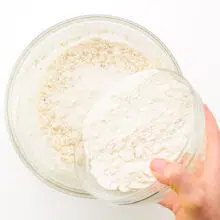 A hand holds a flour mixture, pouring it into a bowl with wet ingredients.