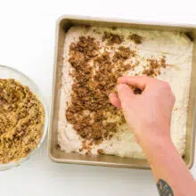 A hand distributes cinnamon filling mixture over cake batter in a baking pan.