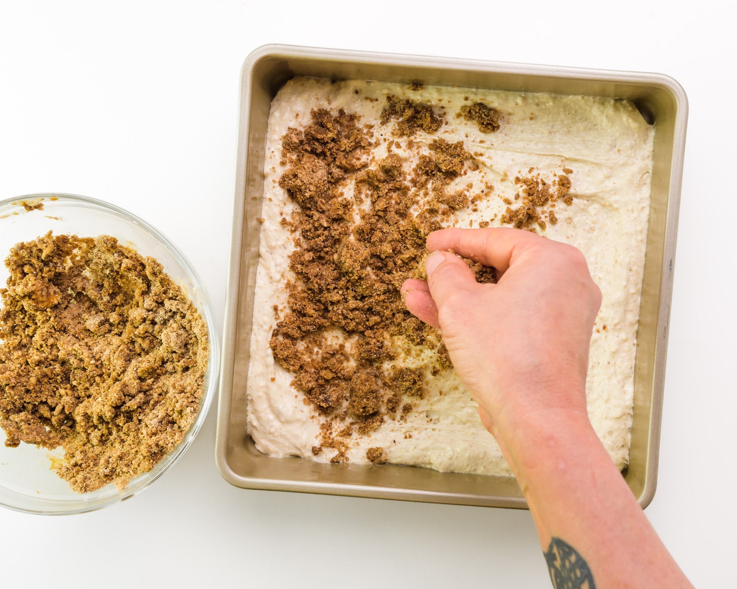 A hand distributes cinnamon filling mixture over cake batter in a baking pan.