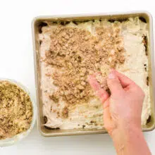 A hand is distributing crumble topping across cake batter in a baking pan.