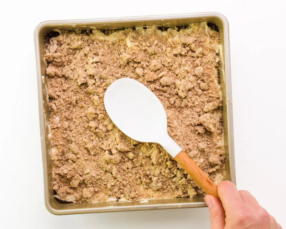 A hand holds a spatula, gently pressing crumble topping into an unbaked vegan coffee cake.