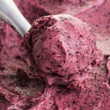 An ice cream scoop is full of blueberry vegan frozen yogurt, sitting on top of a whole bowl of it.