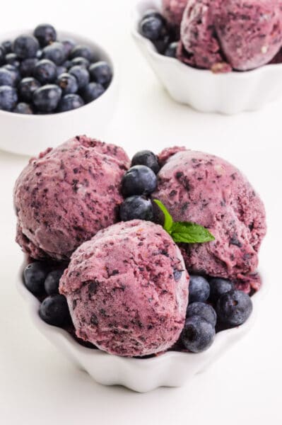 A bowl of blueberry vegan frozen yogurt has fresh blueberries and mint sprigs. There is another bowl in the background next to a bowl of fresh blueberries.