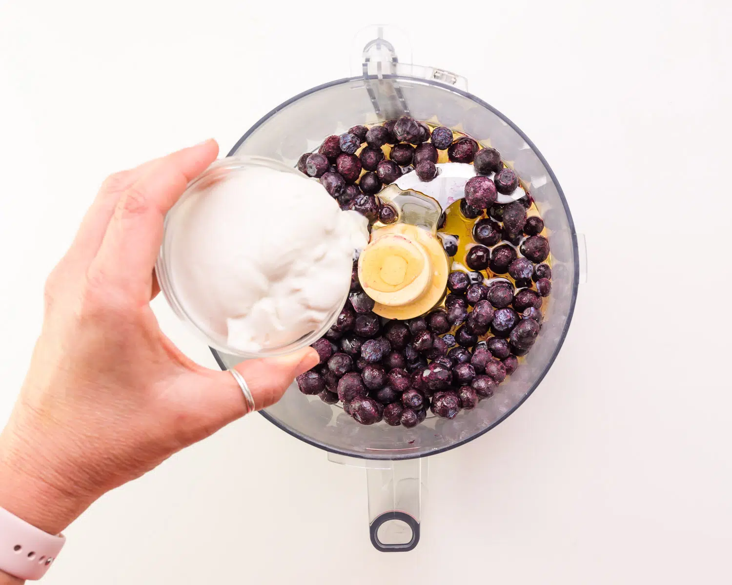 A hand holds a bowl of yogurt, pouring it into a food processor with frozen berries.