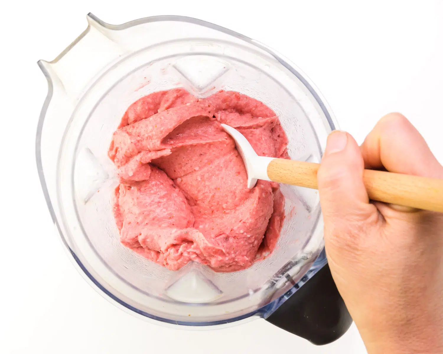 A hand holds a spatula, stirring a frozen strawberry mixture in a blender.