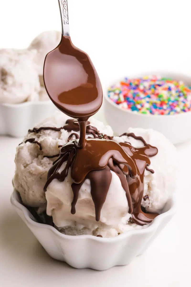 A spoon drizzles chocolate topping on a bowl of ice cream. There's another bowl of ice cream and a bowl of sprinkles in the background.