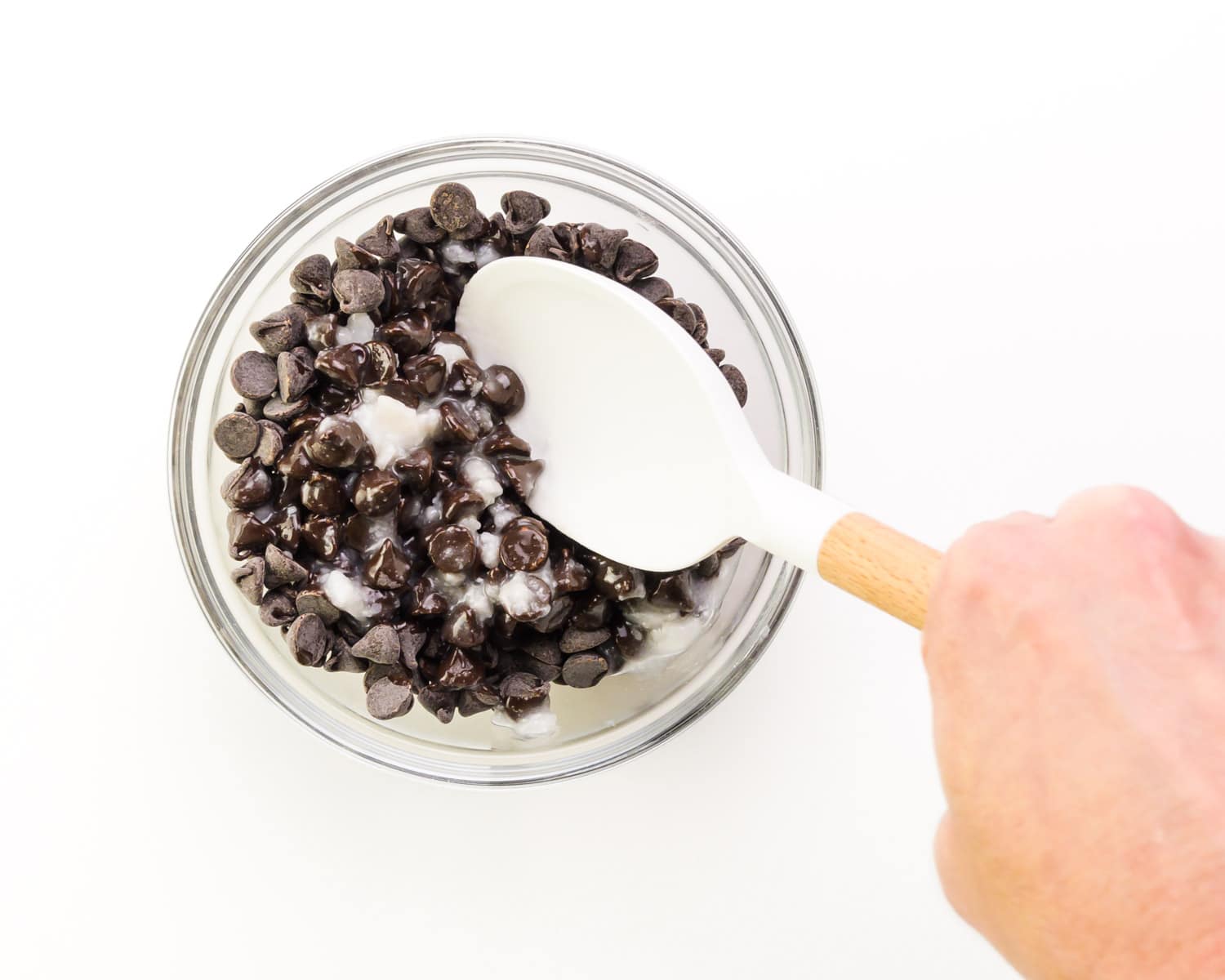 A hand holds a spatula, stirring together chocolate chips and coconut oil.
