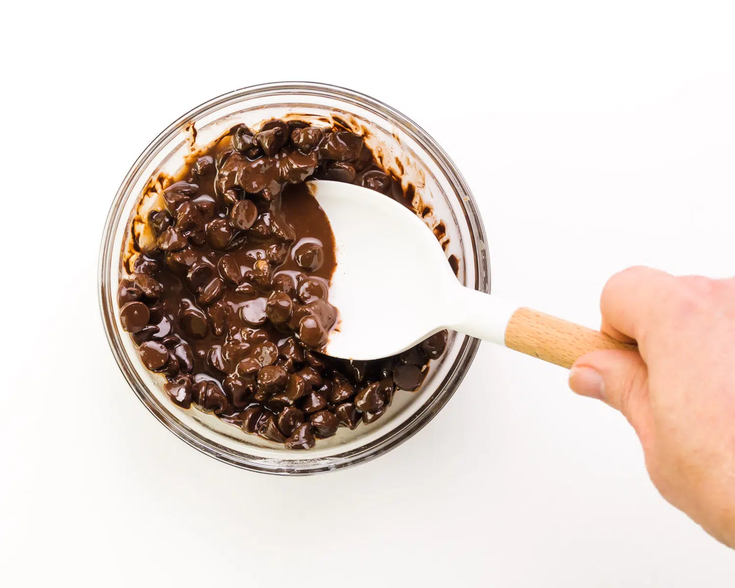 A hand holds a spatula stirring melted chocolate chips in a bowl.