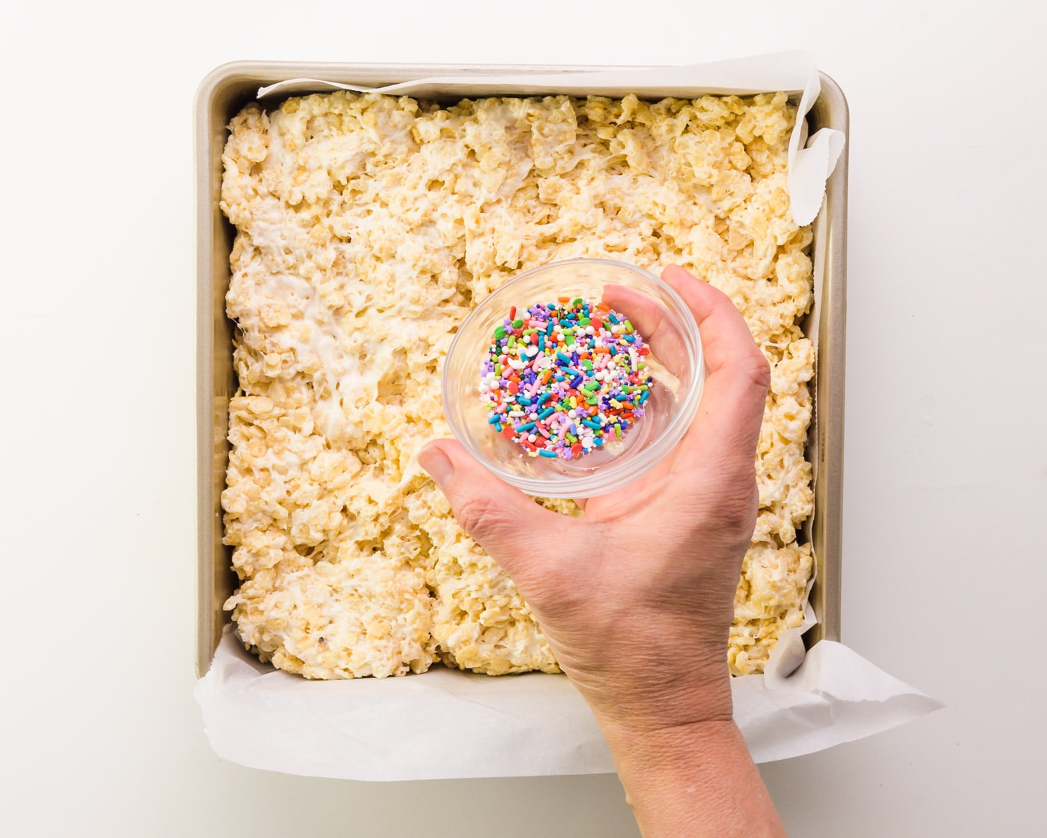 A hand holds a bowl of sprinkles over vegan Rice Krispies in a pan.