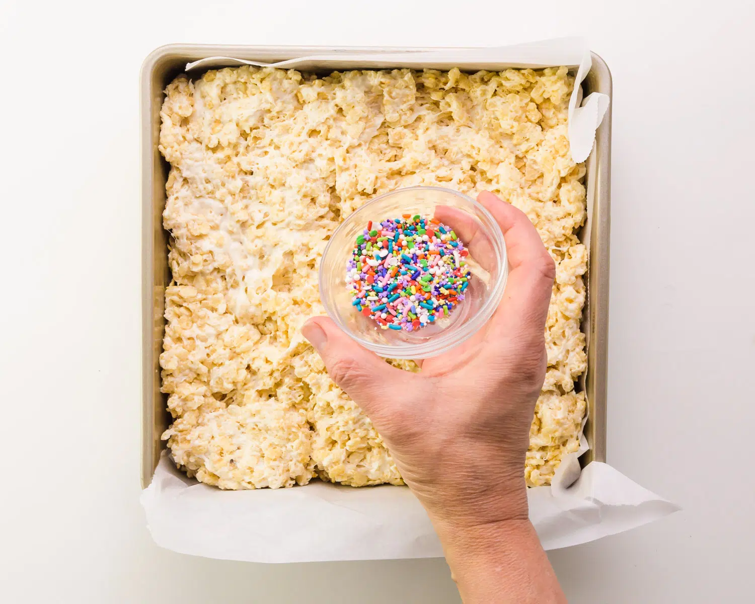 A hand holds a bowl of sprinkles over vegan Rice Krispies in a pan.