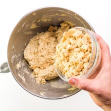 A hand holds a bowl of chopped graham crackers, pouring them into a bowl with cookie batter.