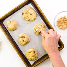 A hand presses marshmallows into cookies. There are bowls of chocolate chips, marshmallows, and chopped graham crackers next to the pan.