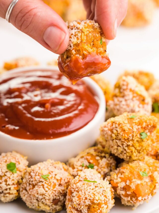 A hand holds a sweet potato tater tot dipped in ketchup, dangling over a plate with more tater tots and a bowl of ketchup.