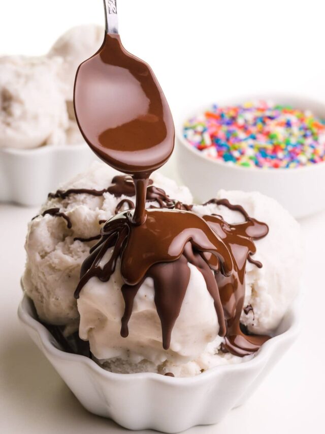 A spoon drizzles chocolate topping on a bowl of ice cream. There's another bowl of ice cream and a bowl of sprinkles in the background.