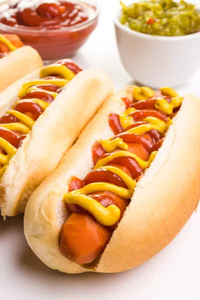 Carrot dogs are topped with ketchup and mustard. There are bowls of ketchup and relish in the background.