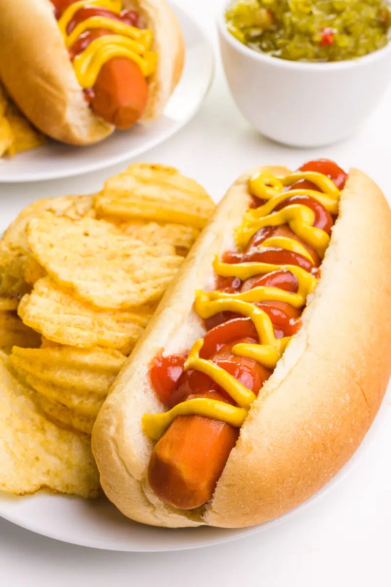 A vegan carrot dog is in a bun on a plate next to potato chips. There's another plateful in the background next to a bowl of relish.
