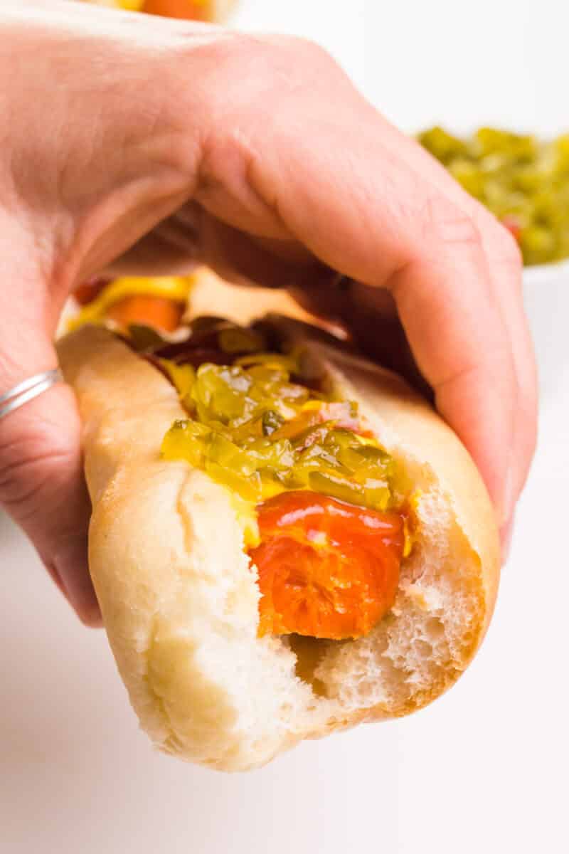 A hand holds a vegan carrot dog in a bun with relish on top.