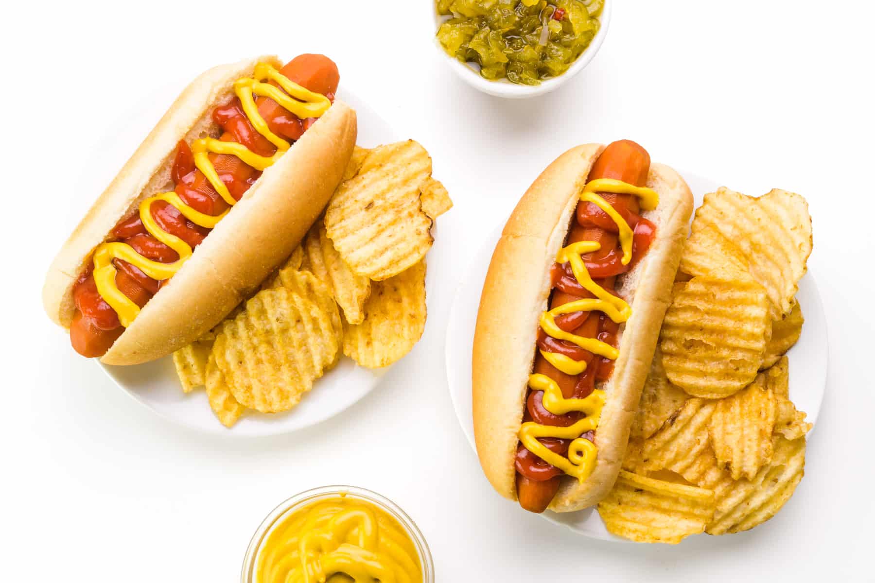 Looking down on plant-based hot dogs on plates sitting next to potato chips. There are bowls of relish and mustard nearby.