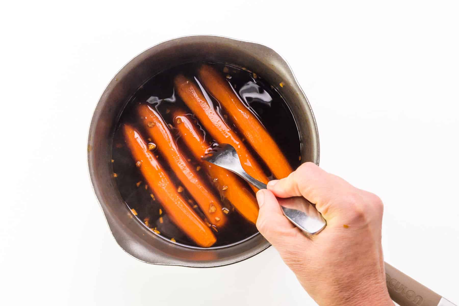 A handholds a fork, pushing it into carrots in a saucepan, testing for doneness.