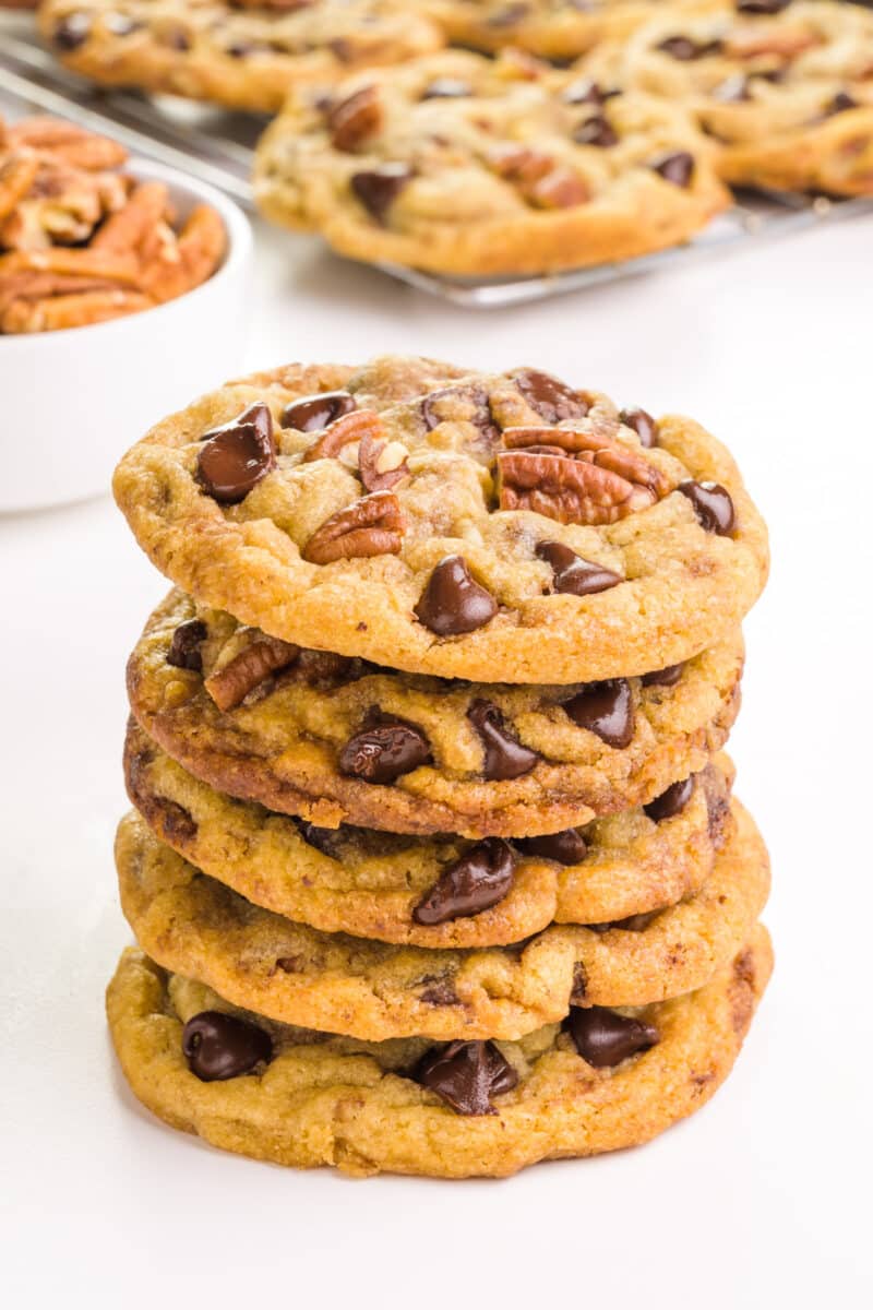 A stack of chocolate chip pecan cookies in front of a bowl of pecans and more cookies in the background.