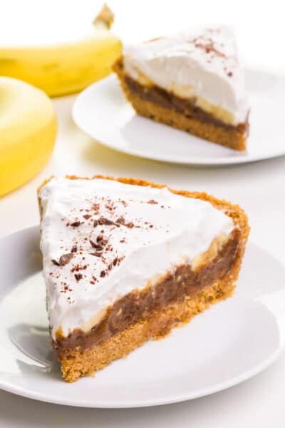 Two slices of vegan banoffee pie sit on separate plates. There are two bananas beside the plates.