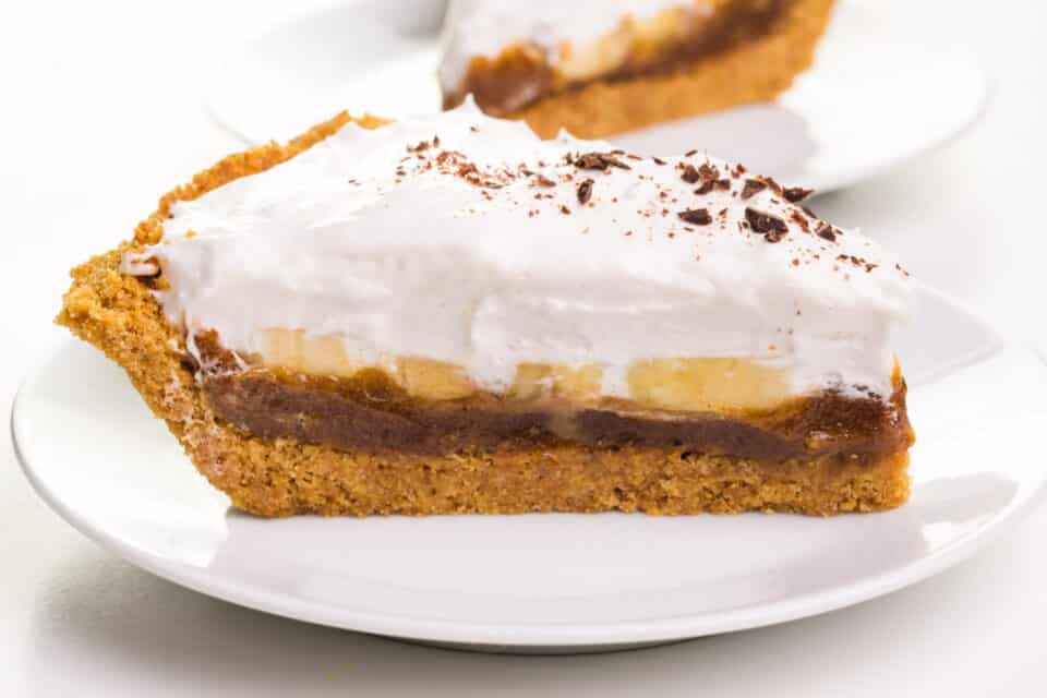 A slice of banoffee pie sits sideways on a plate, showing off the layers. Another slice is in the background.