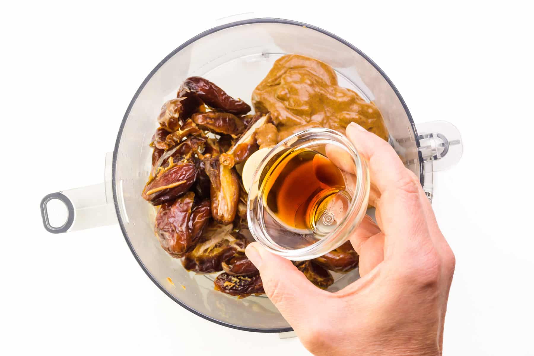 A hand holds a bowl of maple syrup, adding it to a food processor with dates and nut butter.