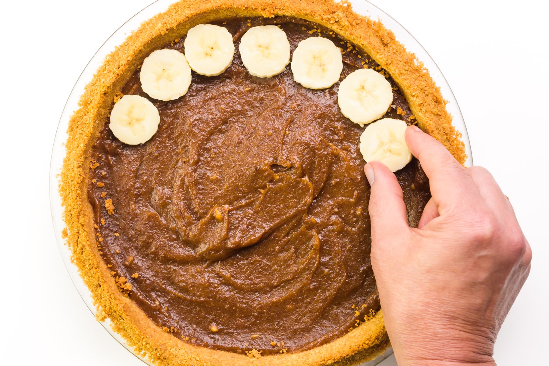 A hand places banana slices over filling in a pie crust.