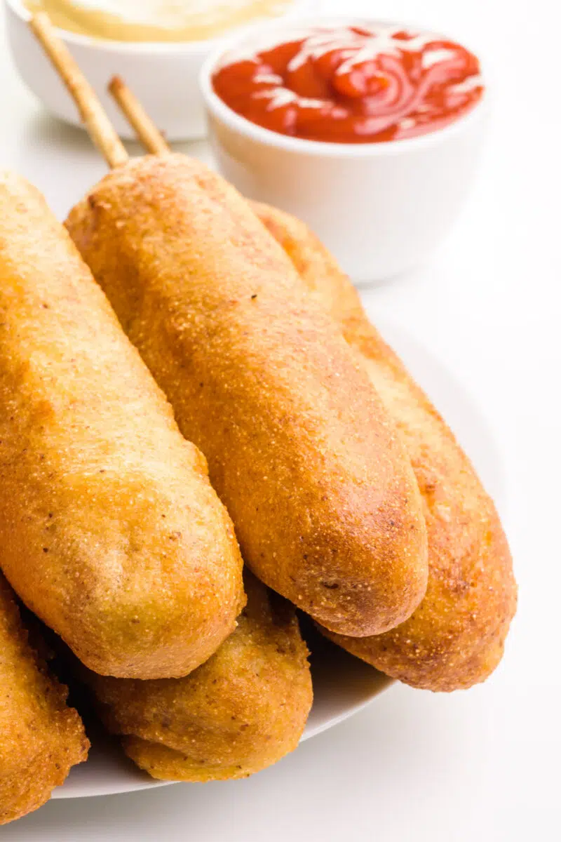 Vegan corn dogs are stacked on a plate, sitting in front of bowls of ketchup and mustard.