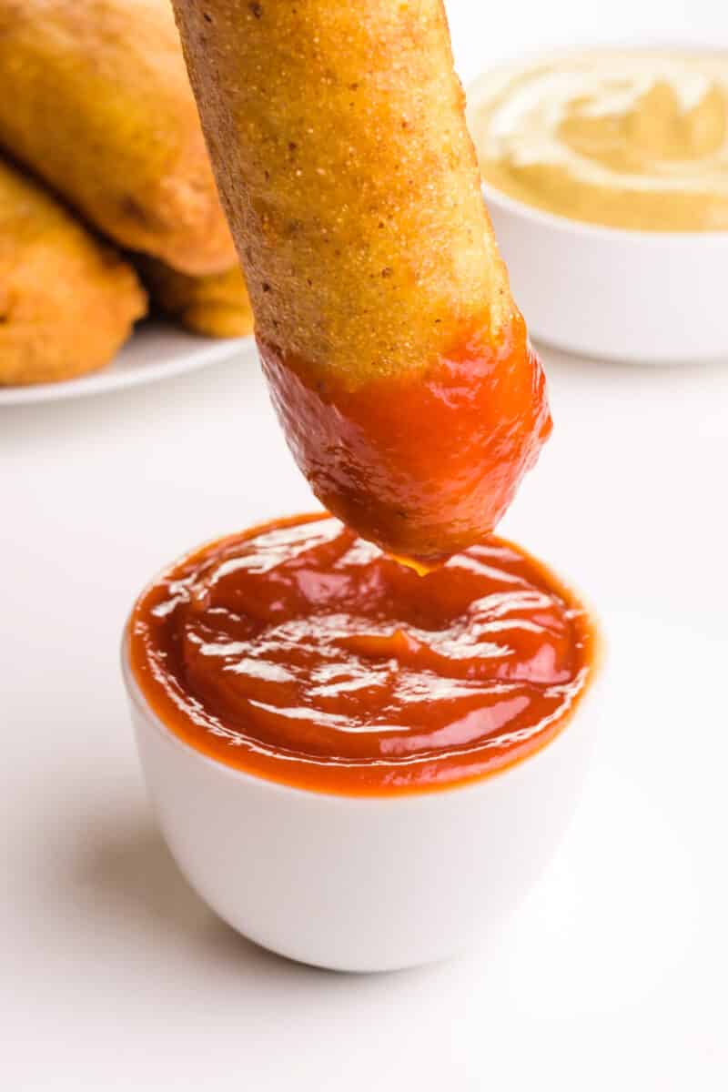 A corn dog is being dipped in ketchup. It sits in front of a bowl of mustard and more corn dogs in the background.