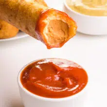 A vegan corn dog has a bite taken out of it and is hovering over a bowl of ketchup. There's a bowl of mustard in the background.