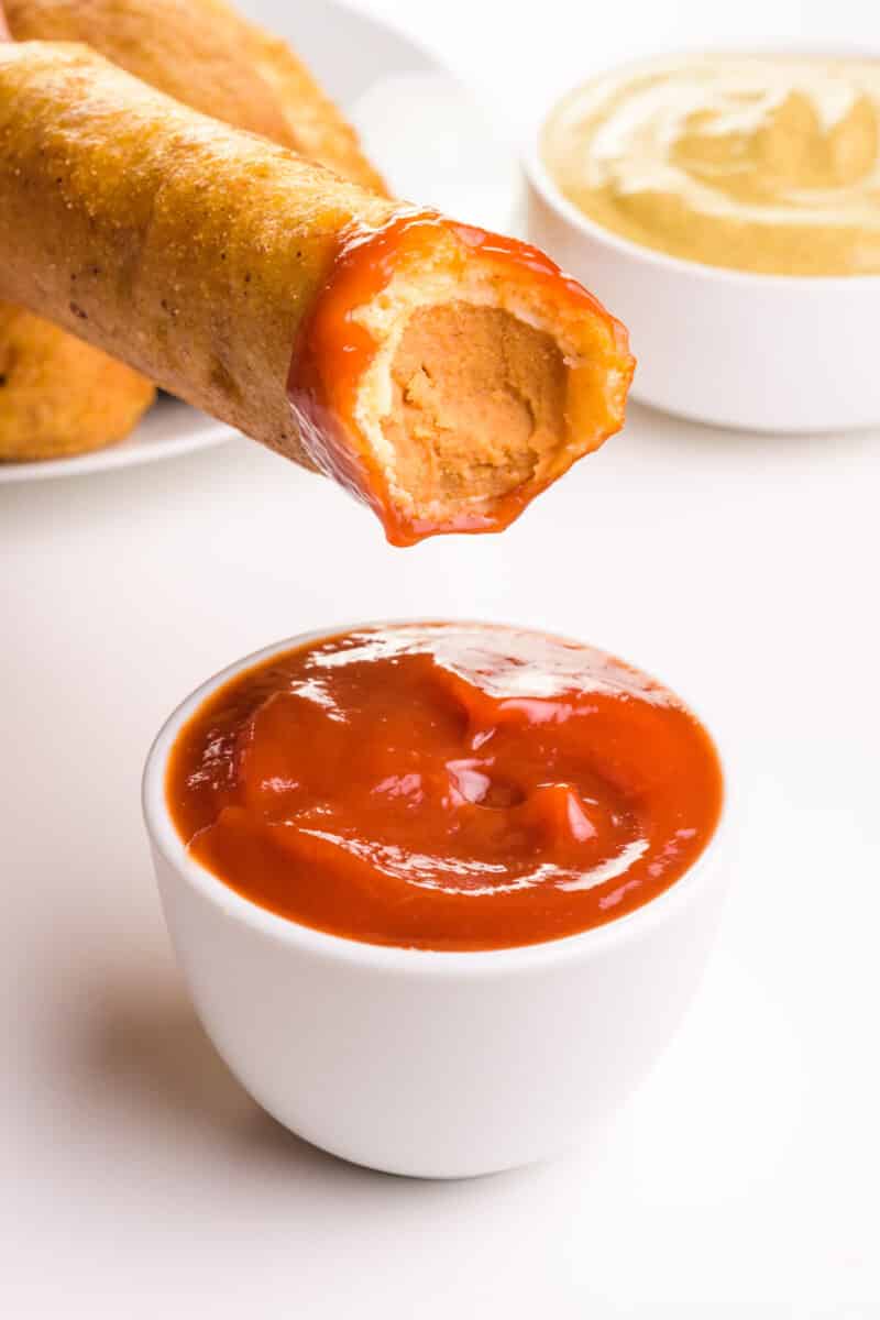 A vegan corndog has a bite taken out of it and is hovering over a bowl of ketchup. There is a bowl of mustard in the background.