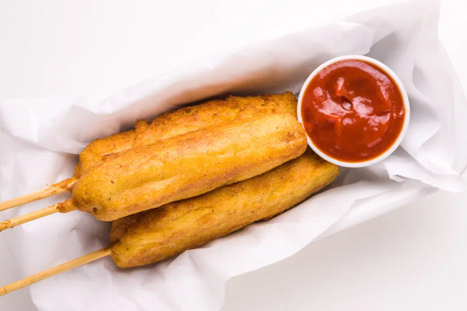 Looking down on vegan corn dogs in a basket sitting next to a bowl of ketchup.