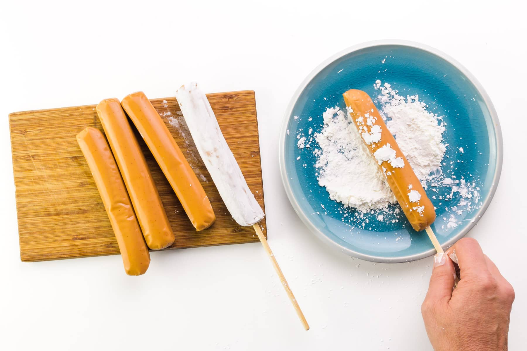 A hand holds a hot dog on a stick, rolling in on a plate with cornstarch. There are more hot dogs on a wooden cutting board by the plate.