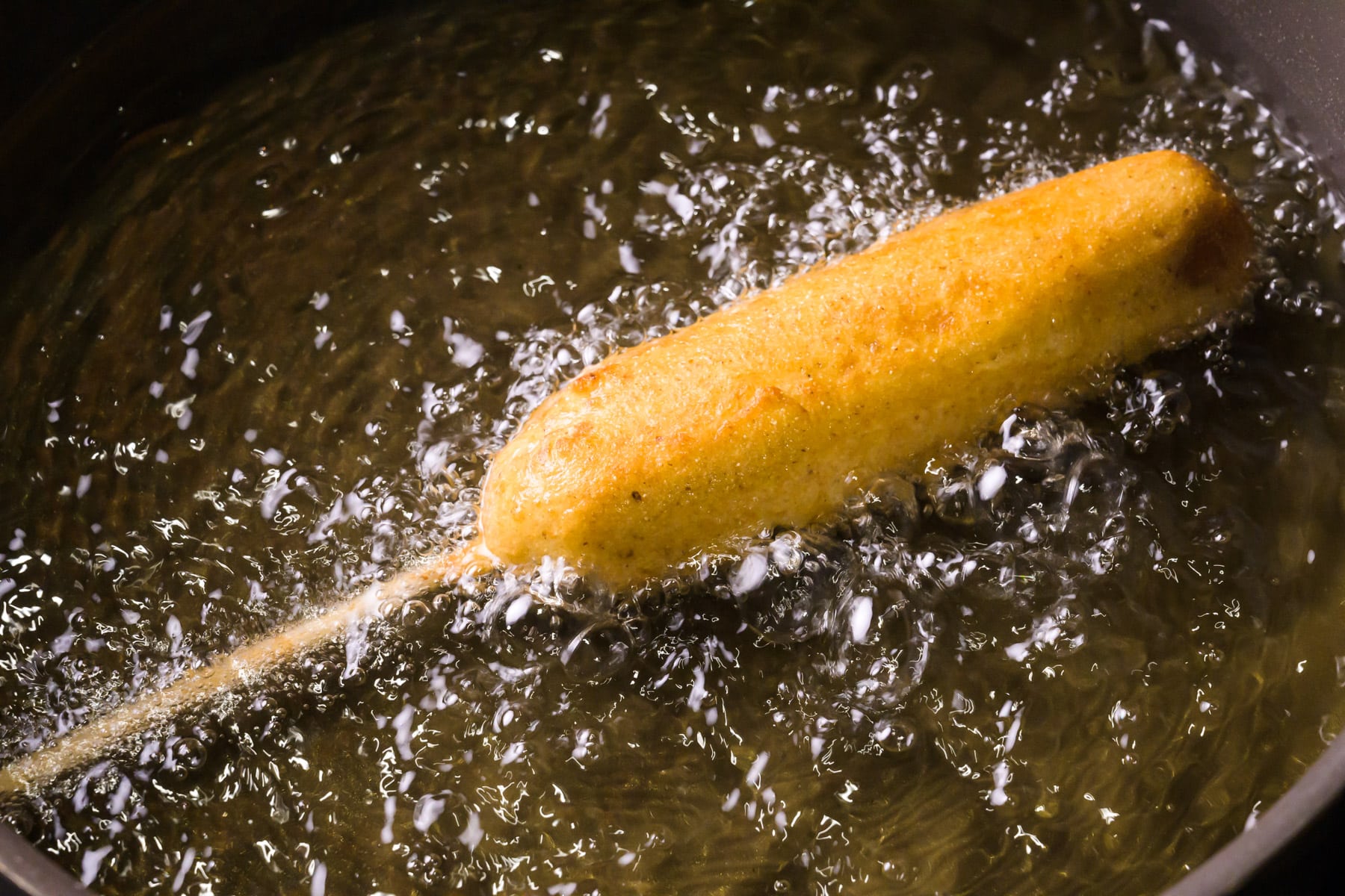 A corn dog is frying in hot oil in a skillet.